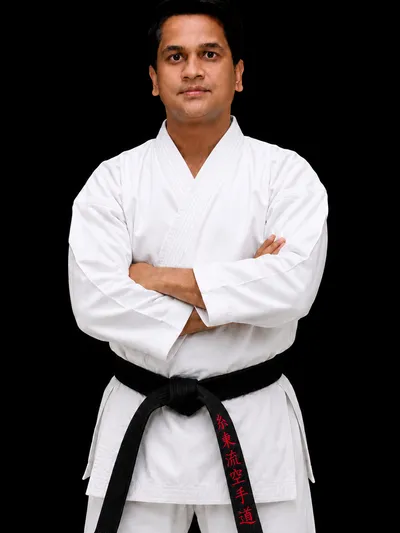 A studio portrait of Sensei Amit More wearing a traditional white karate gi with a frayed black belt